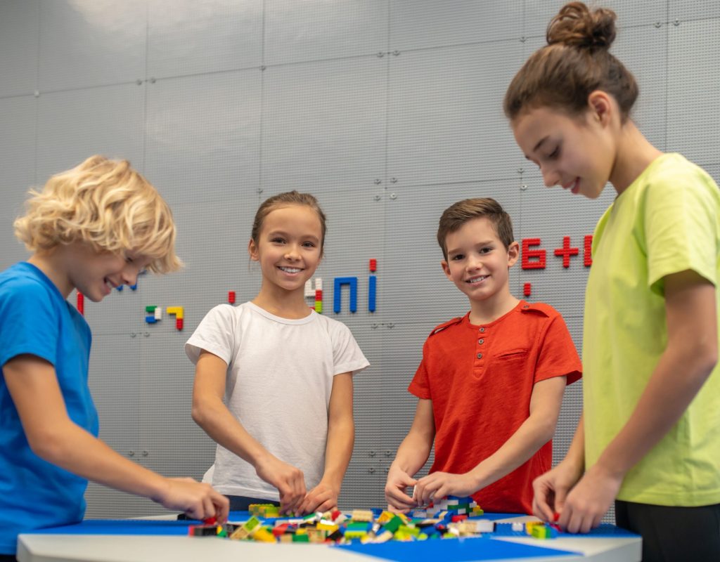 Good moments. Boy and girl smiling at camera playing with friends constructor standing near table in indoor play area